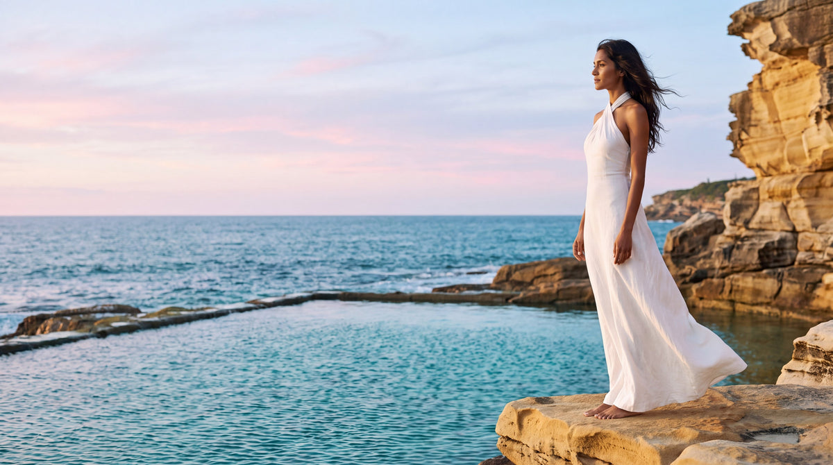 Woman in a white halter maxi dress standing on sandstone rocks by an ocean pool at sunset
